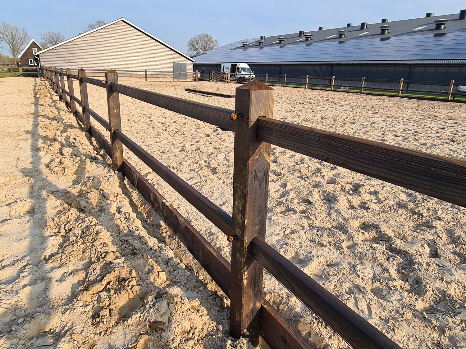 Een rijbak wordt begrensd door een elegant paardenhek. Op de achtergrond staan verschillende stalgebouwen.