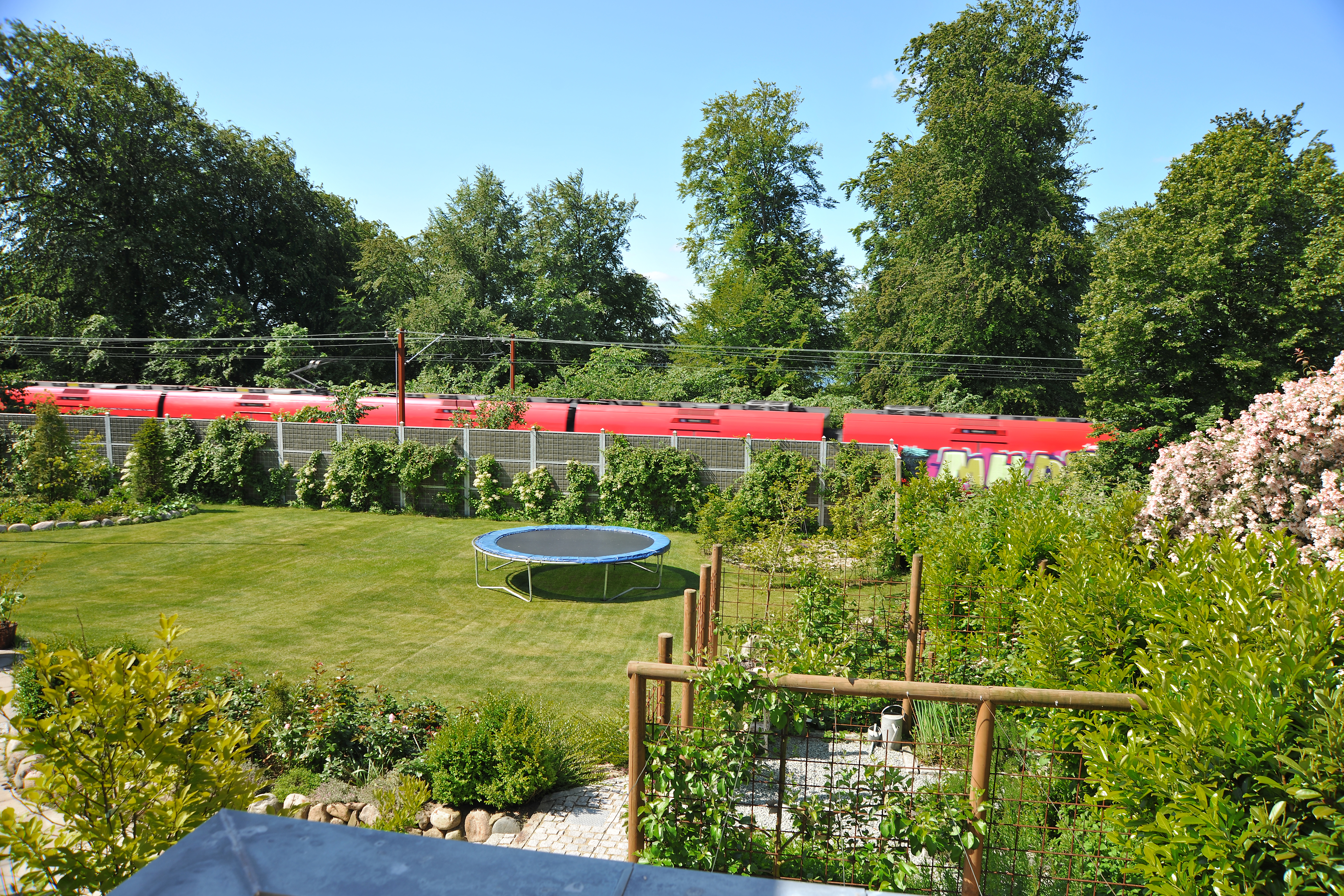 Een Noistop® Steel geluidswerende schutting van Poda Omheiningen met begroeiing geïnstalleerd in een tuin om geluidsoverlast van een spoorlijn vlak naast de tuin te weren.
