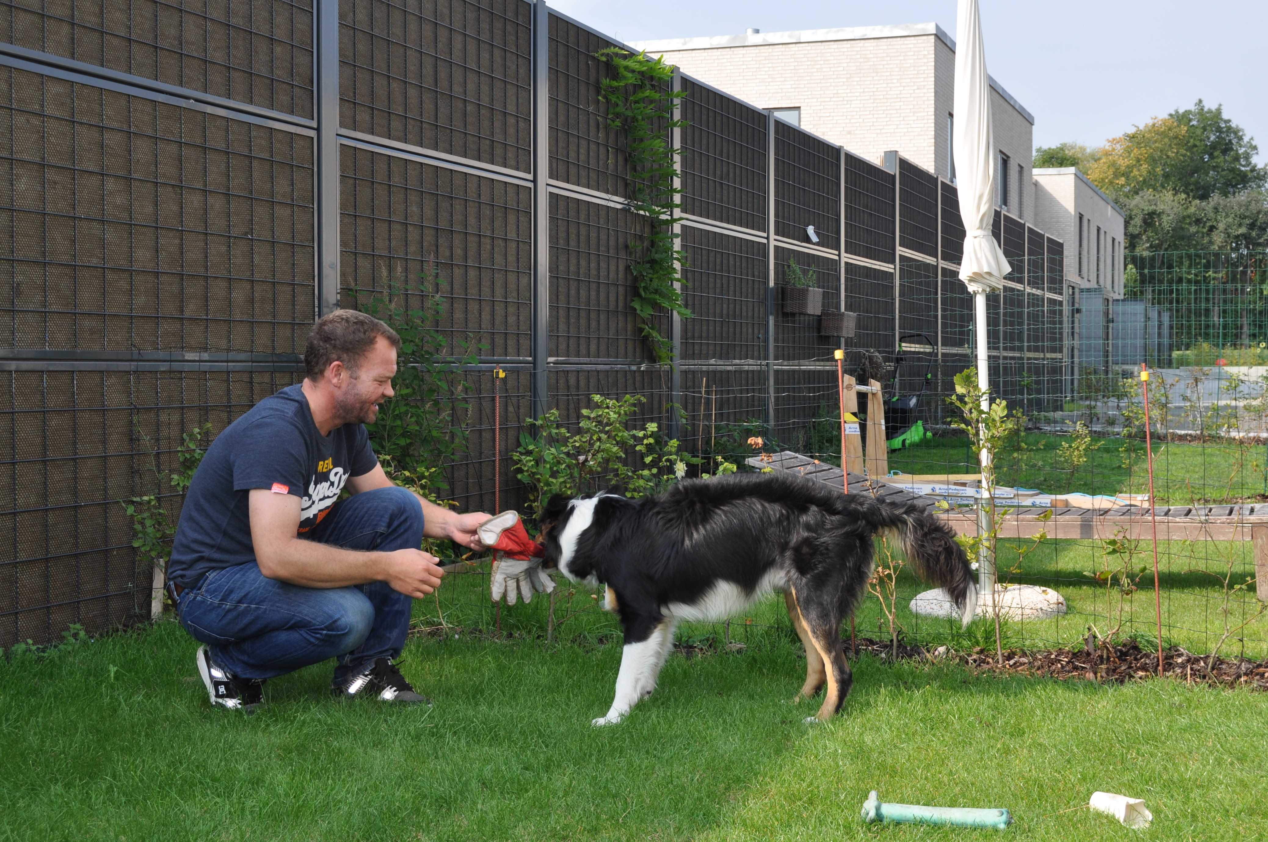 Een man en zijn hond zitten voor een Noistop® Steel geluidswerende schutting van Poda Omheiningen dat in een tuin is geplaatst om burenoverlast te weren.