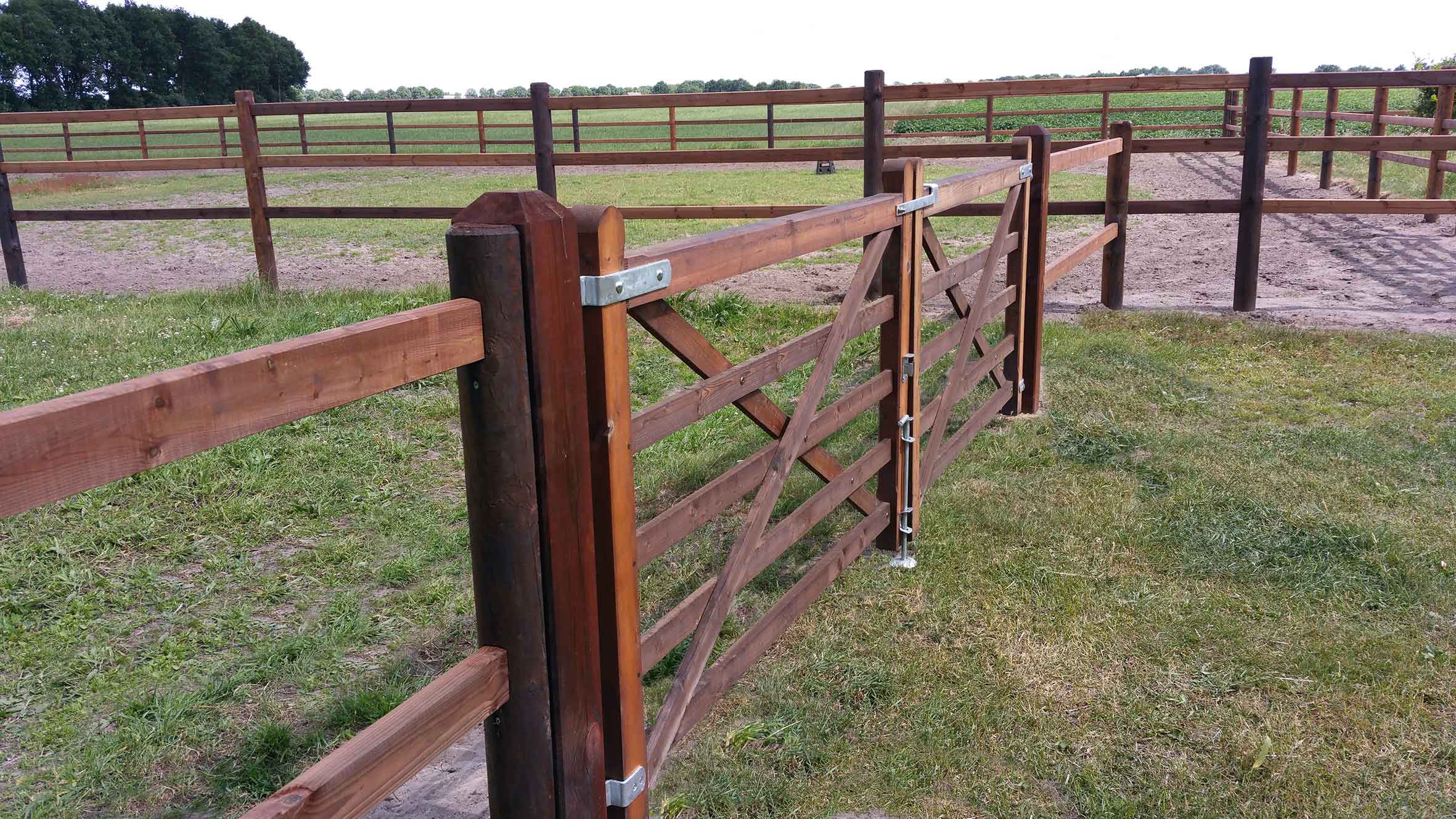 Een paardenomheining scheidt twee paddocks van elkaar. Een dubbele houten poort geeft toegang tot een paddock.